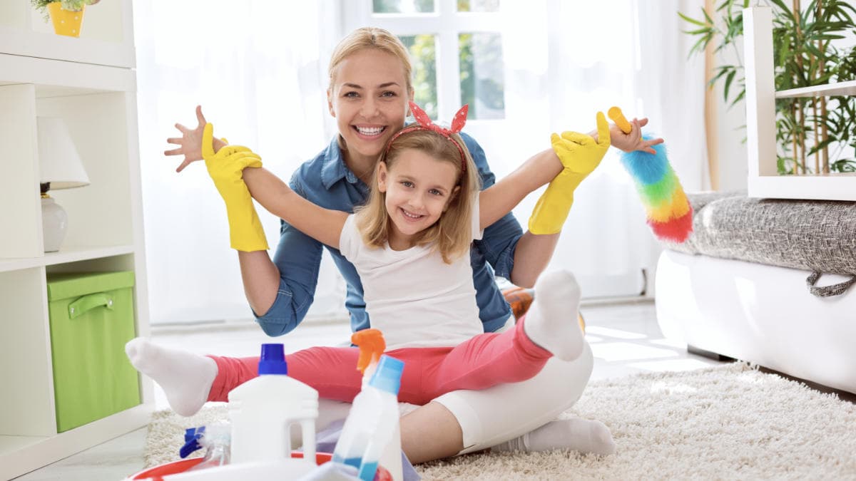 Smiling woman and child in yellow gloves, sitting on a carpet with cleaning supplies, symbolizing joyful house cleaning.