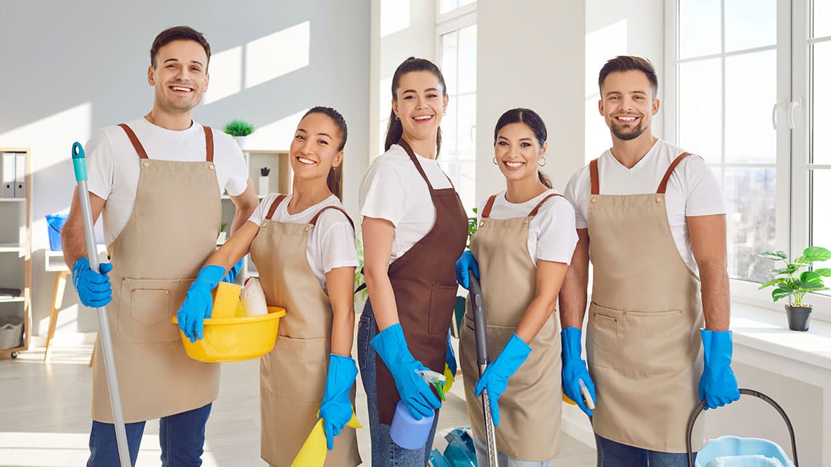 Smiling house cleaning team in aprons and gloves standing in a bright home, holding cleaning tools and supplies.