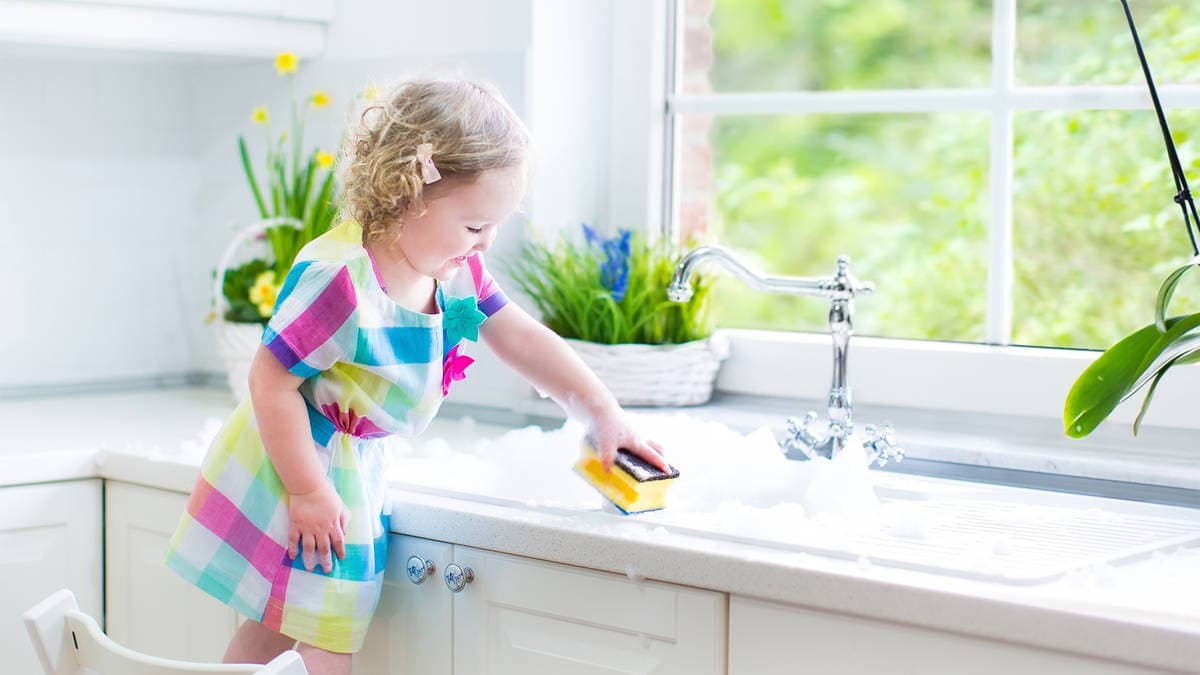 A child in a colorful dress scrubs a sink filled with soap suds, surrounded by plants and sunlight in a bright kitchen.