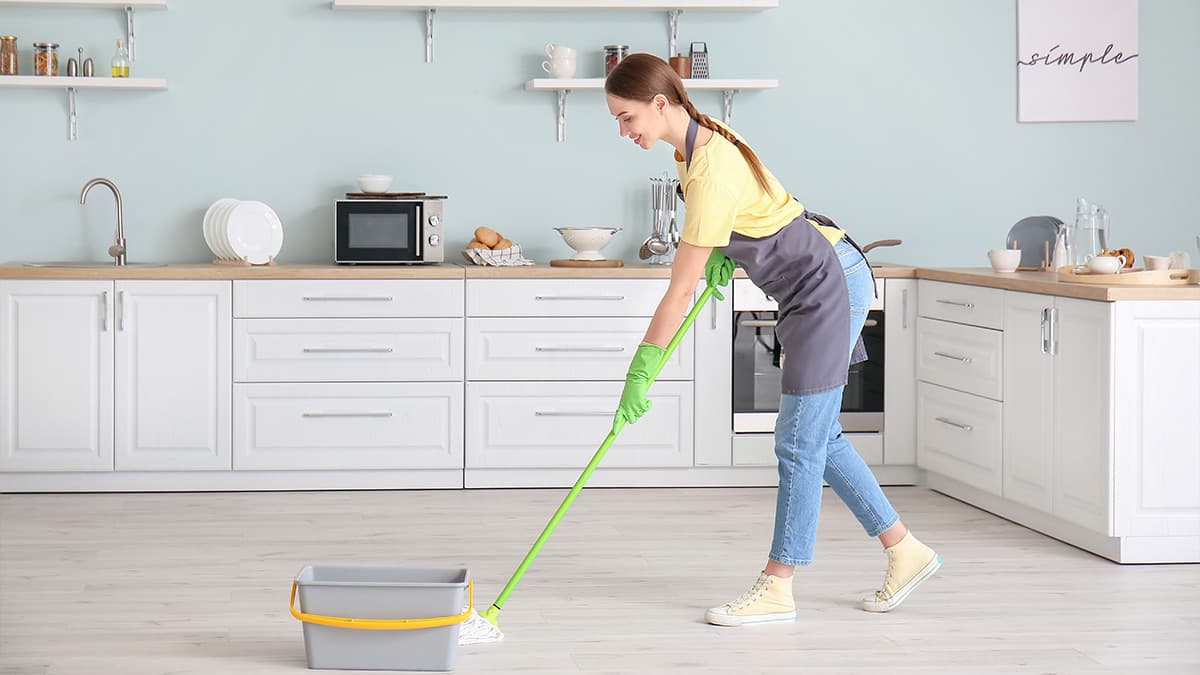 A woman cleaning a modern kitchen floor with a mop, wearing gloves and an apron, showcasing cleanliness and organization.