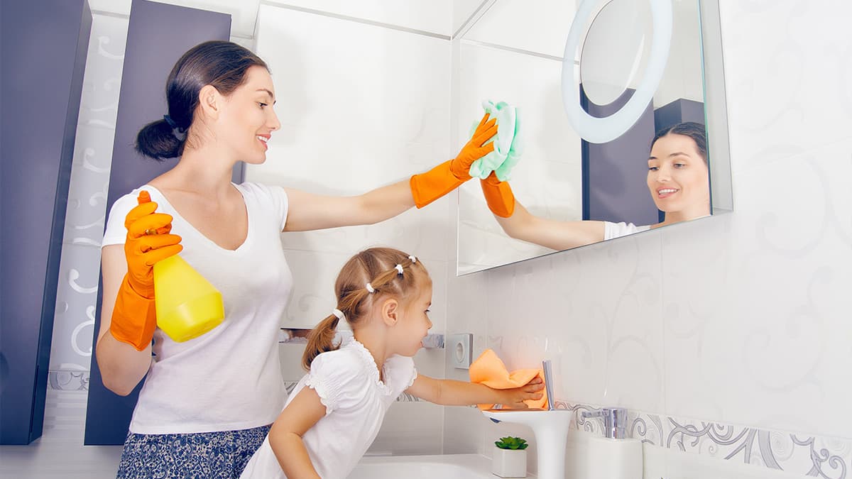A woman and a child clean a bathroom together, using spray cleaner and cloths, showcasing family involvement in cleaning tasks.