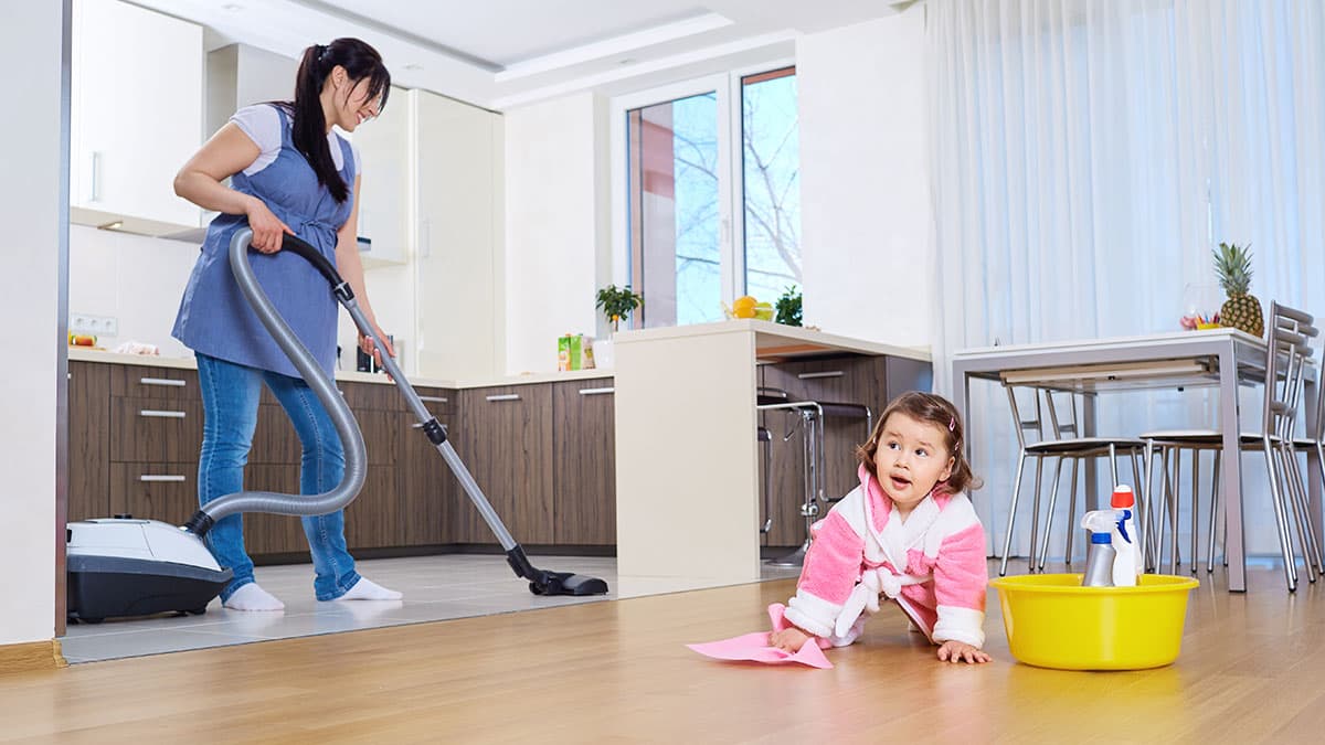 Woman vacuuming the kitchen floor while a young child helps with house cleaning